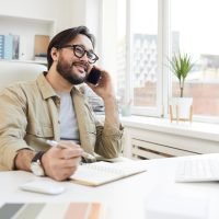 Cheerful communicative young Asian man with beard sitting at table in office and calling customer on phone